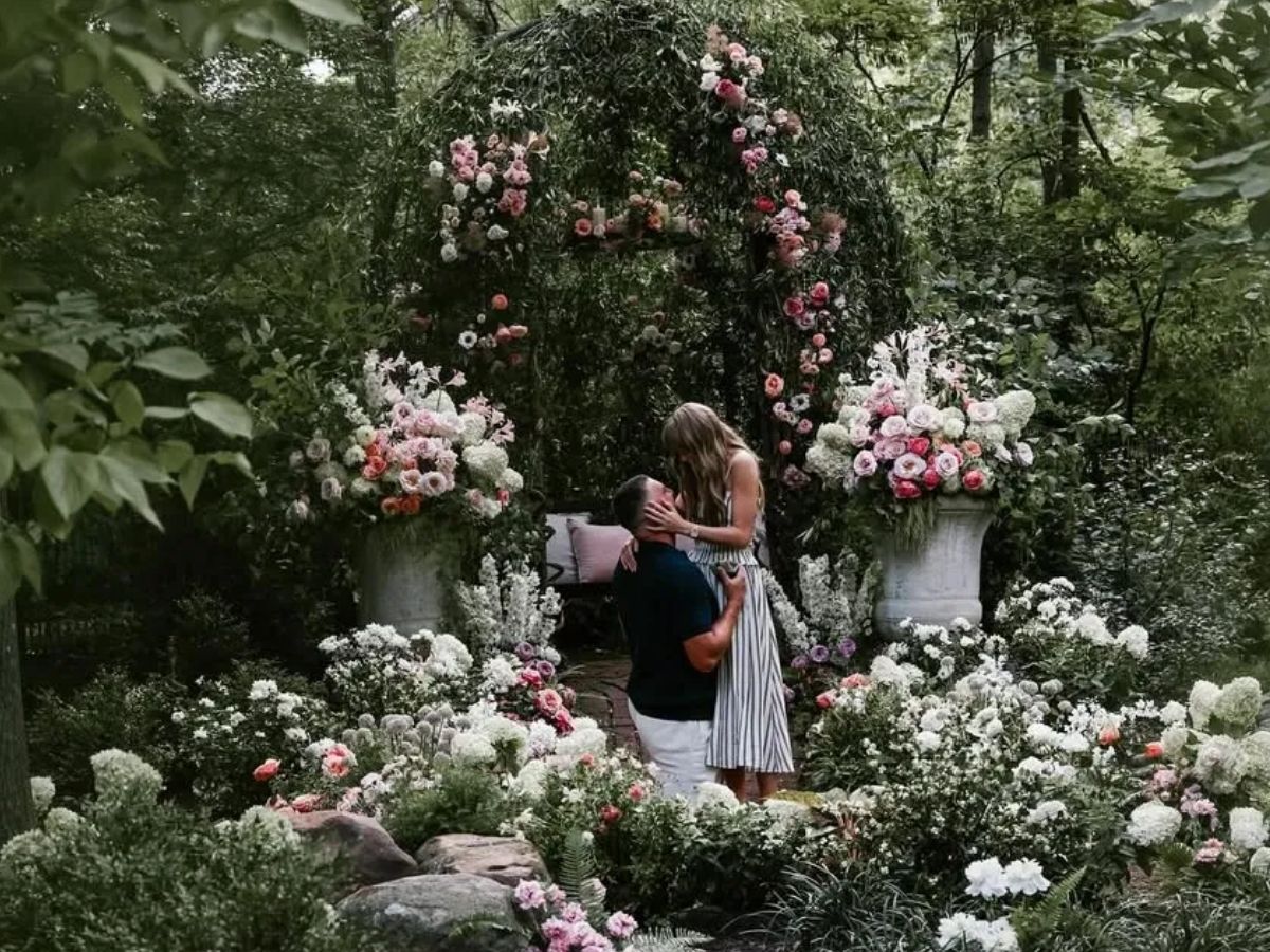 Couple in a proposal scene, surrounded by roses and white blooms in a garden.