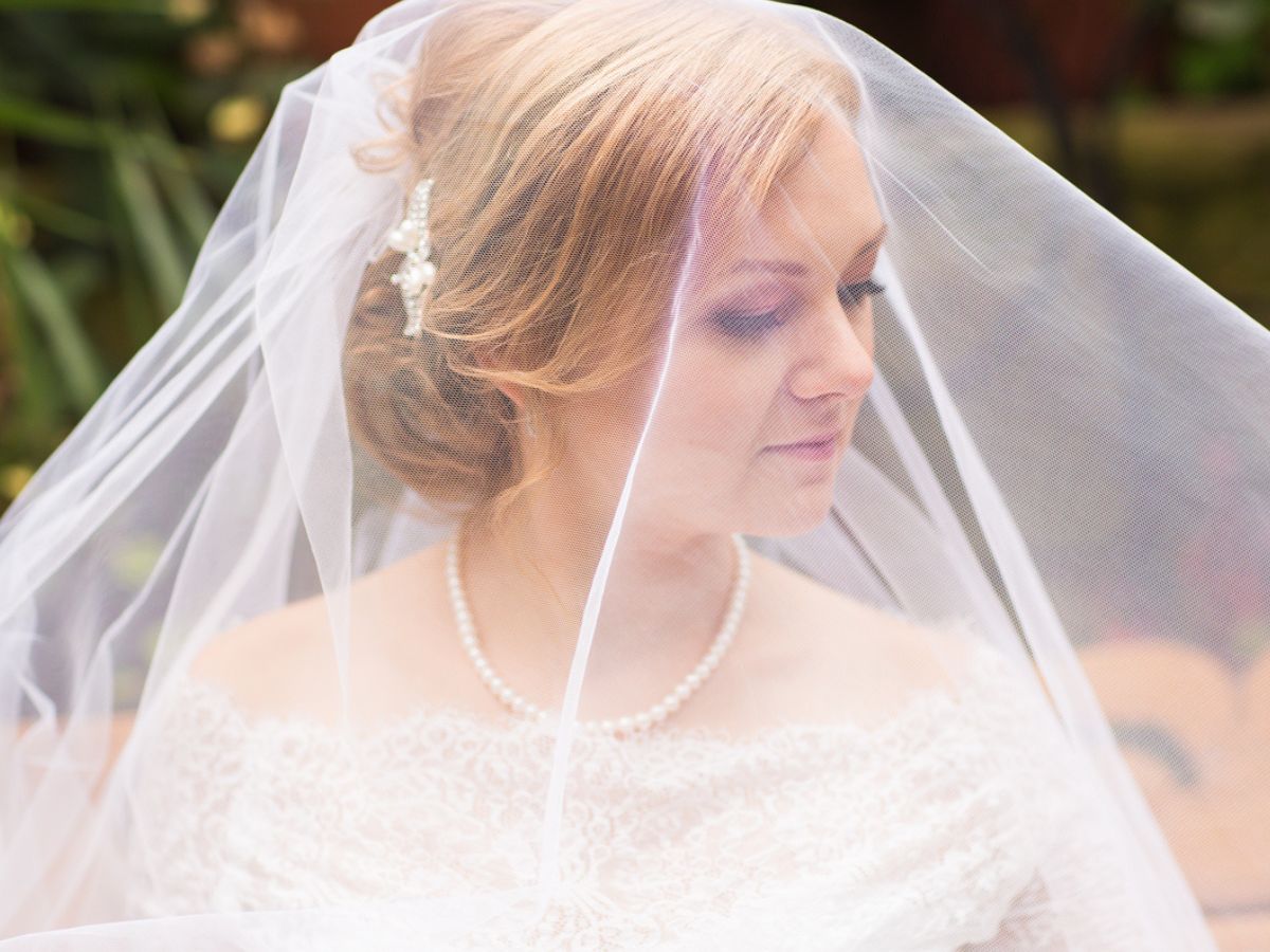Bride in lace dress with veil and pearl necklace, soft natural background.