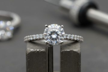 A close-up of a diamond engagement ring with a secure gemstone setting, held in a jeweler's clamp, with tools in the background.