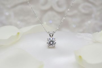 A beautiful solitaire diamond pendant on a silver chain resting on white satin surrounded by white flower petals.