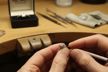 A jeweler's hands carefully inspect and work on a diamond ring with various tools on a wooden workbench.