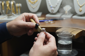 A jeweler cleaning a diamond ring with a small brush over a workbench, with other jewelry in the background.