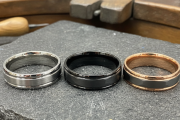 Three tungsten wedding bands in different finishes (silver, black, rose gold tone) resting on a jeweler's workbench.